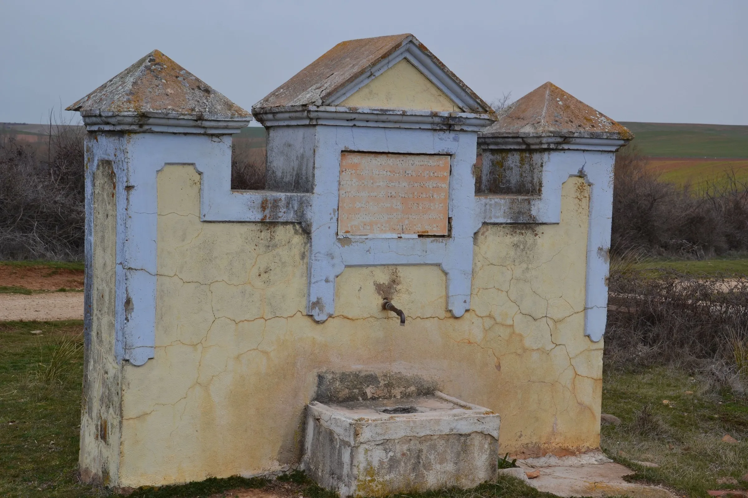 La fuente de Valvieja: un frontón encalado en amarillo y azul, con tres remates piramidales, un caño de hierro y un pilón de piedra al pie