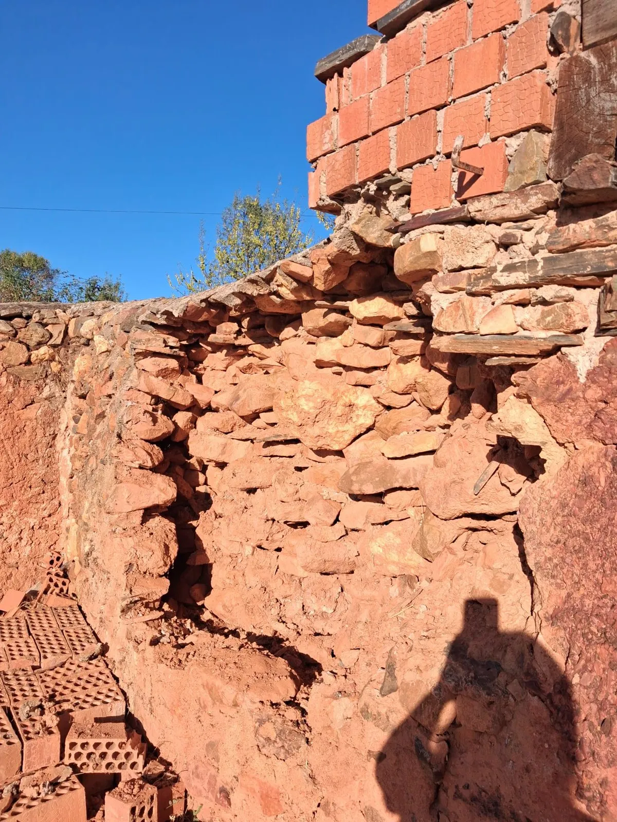 Un tramo de la tapia del cementerio caído, con la piedra roja suelta, el hueco abierto y ladrillos apilados al pie