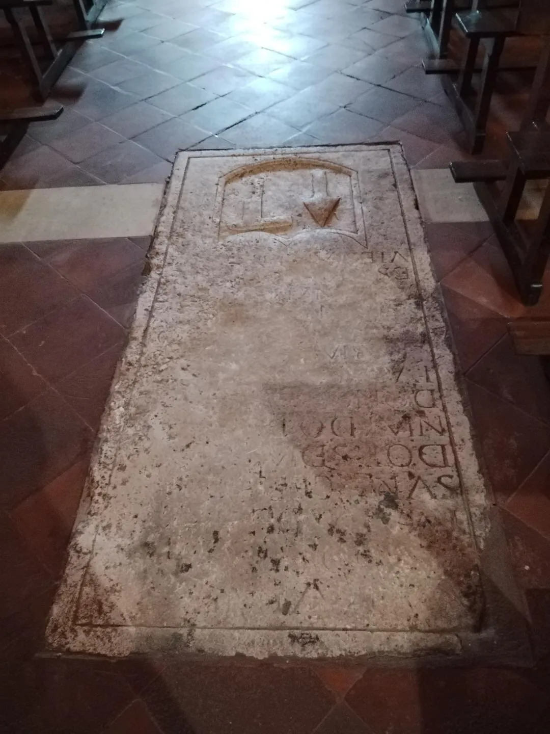 Una losa de piedra clara en el suelo de baldosas de la nave, con un emblema en un recuadro en la cabecera y restos de una inscripción grabada