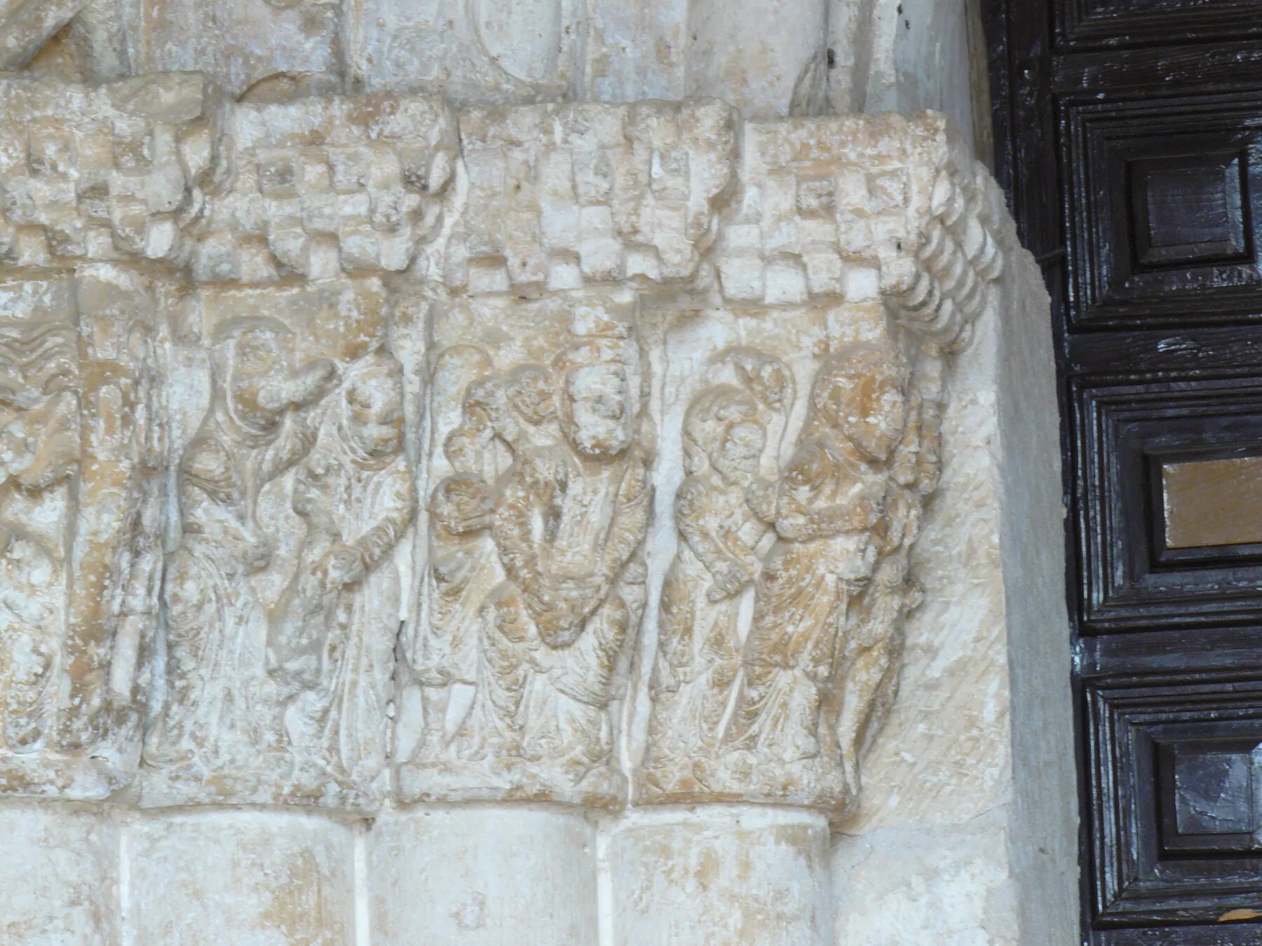 Capiteles románicos junto a la puerta de madera, con grupos de figuras de pie bajo un cimacio de ajedrezado