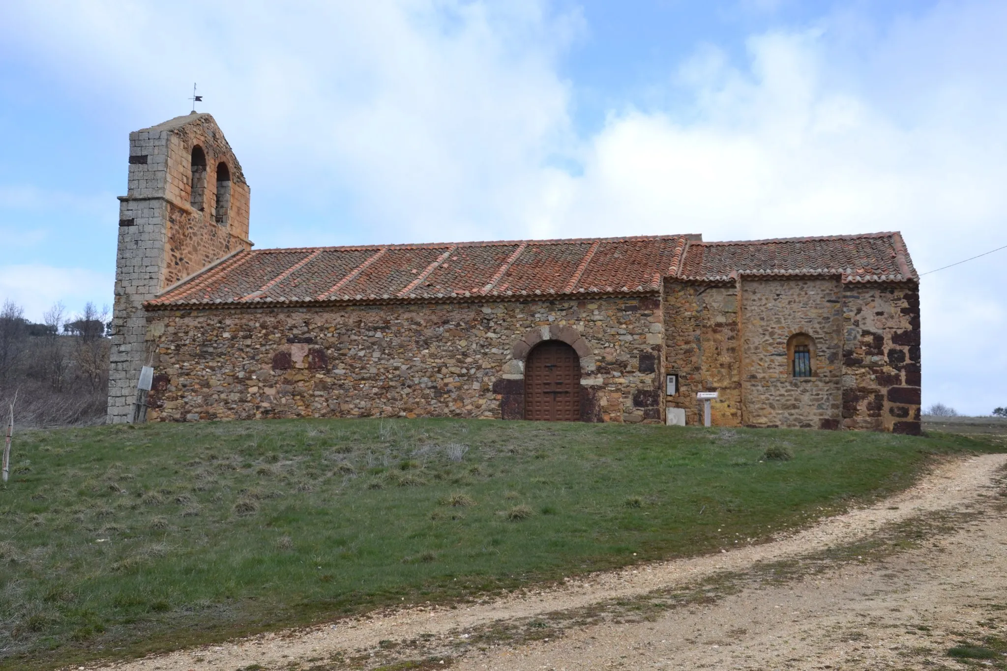 La iglesia de San Pedro de Alquité desde el sur, con la espadaña de dos cuerpos a la izquierda, el atrio de arco de medio punto que protege la portada, y el camino de tierra que sube hasta ella entre los campos