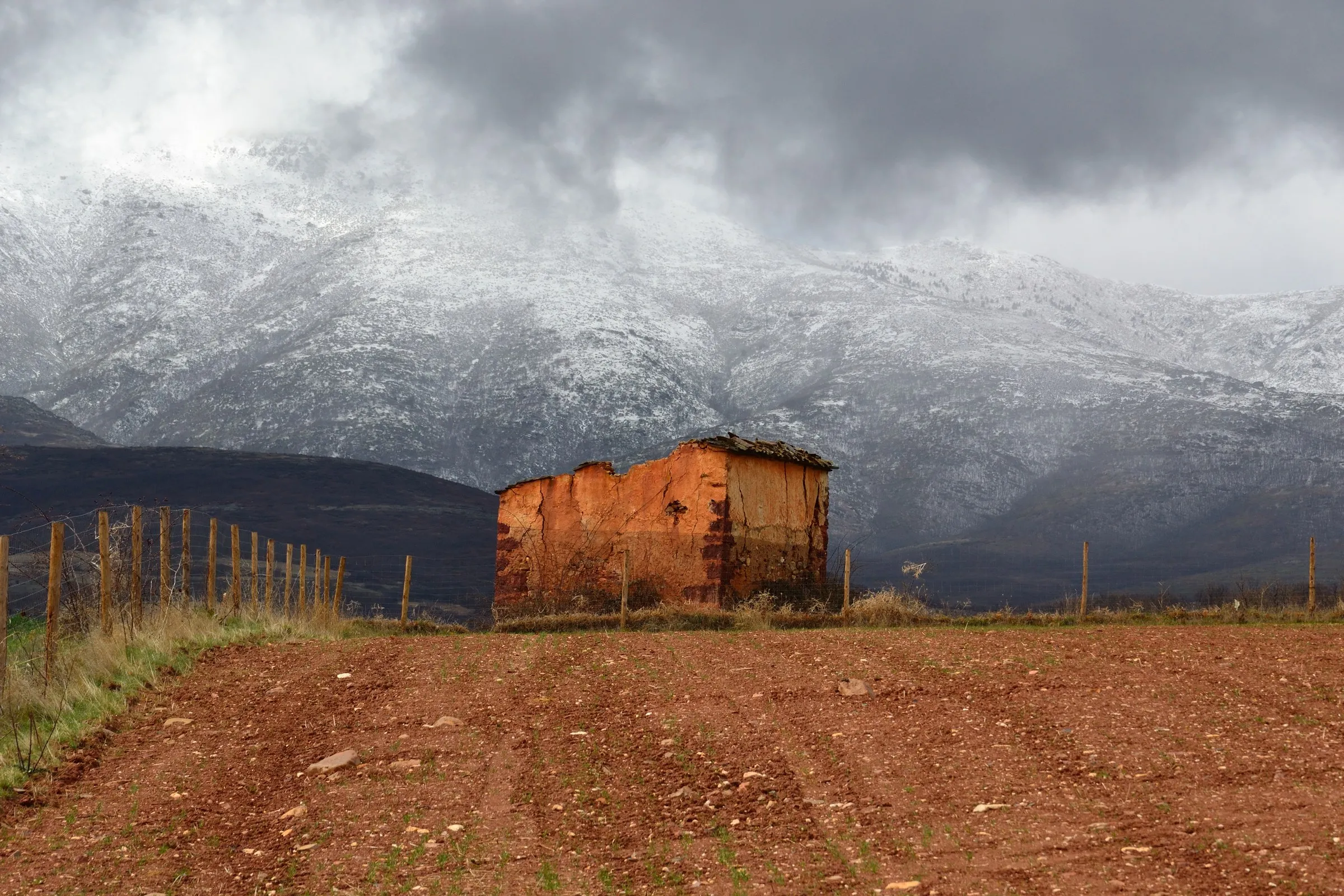 Una caseta rectangular de tapial rojo, con el tejado de lajas medio caído, en un campo arado y con la sierra nevada al fondo