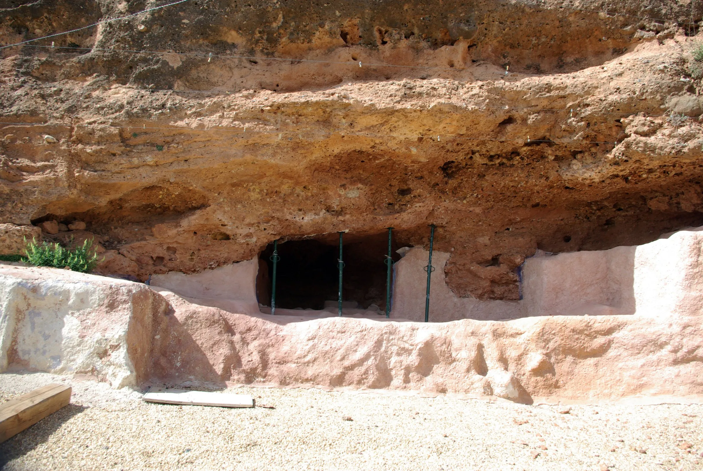 El abrigo de La Peña, una visera de roca anaranjada sobre la boca de una pequeña cueva cerrada con barrotes, con el suelo excavado y protegido