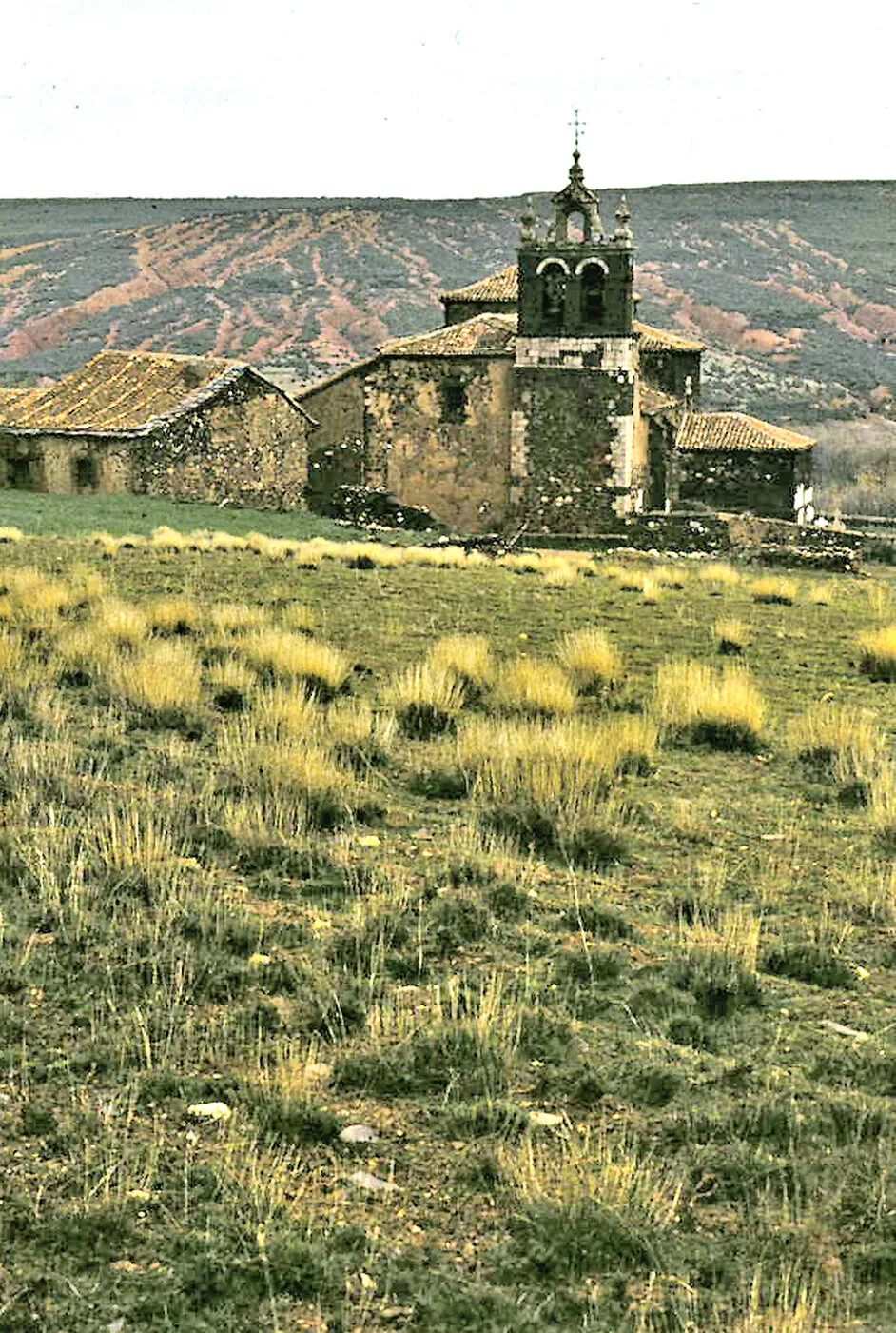 La iglesia de Madriguera y las casas de su alrededor, con un prado delante y los cerros de tierra roja detrás, en 1974