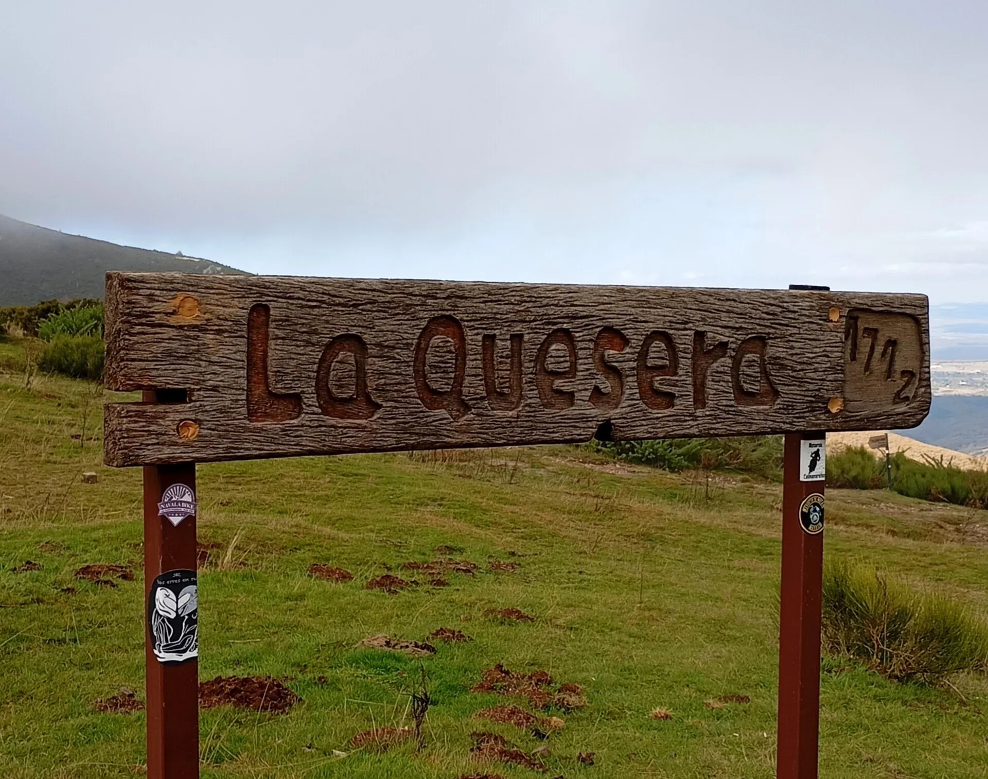El cartel de madera del puerto de la Quesera, con el nombre y la cota talladas, y la sierra al fondo