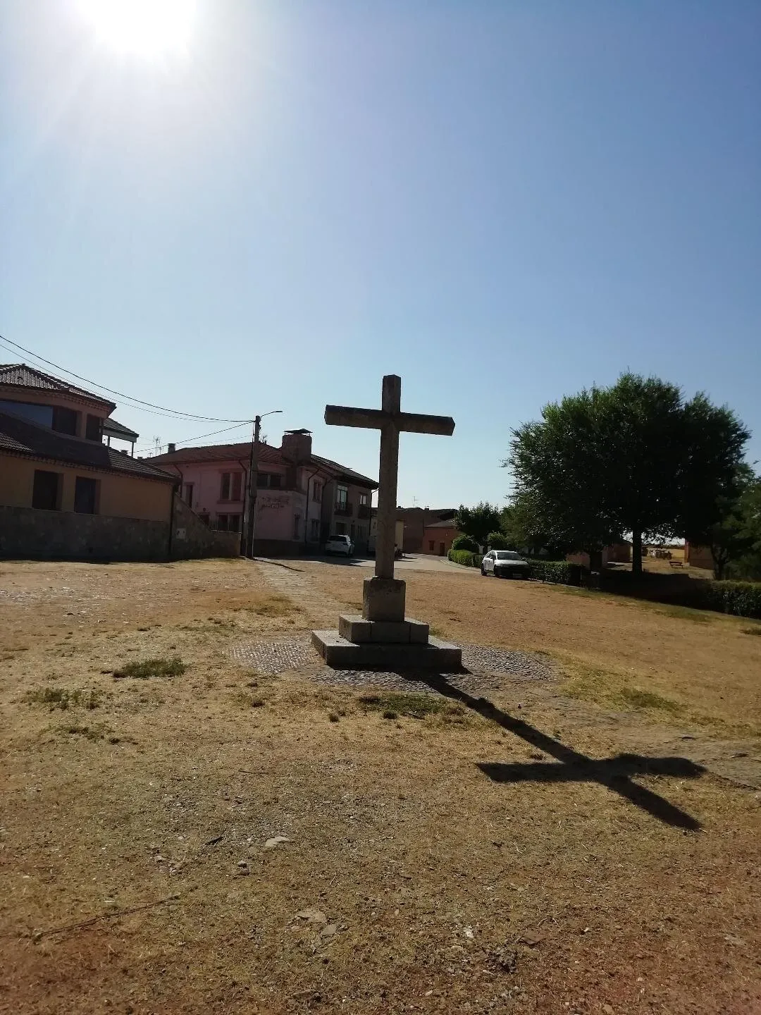 Un crucero de piedra sobre su pedestal en una explanada de hierba seca, con la sombra de la cruz alargada en el suelo
