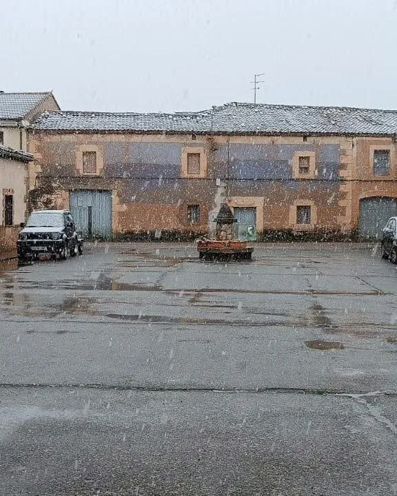 Una plaza asfaltada con una fuente de piedra en el centro y una casona de piedra rojiza con recercados claros detrás, mientras nieva